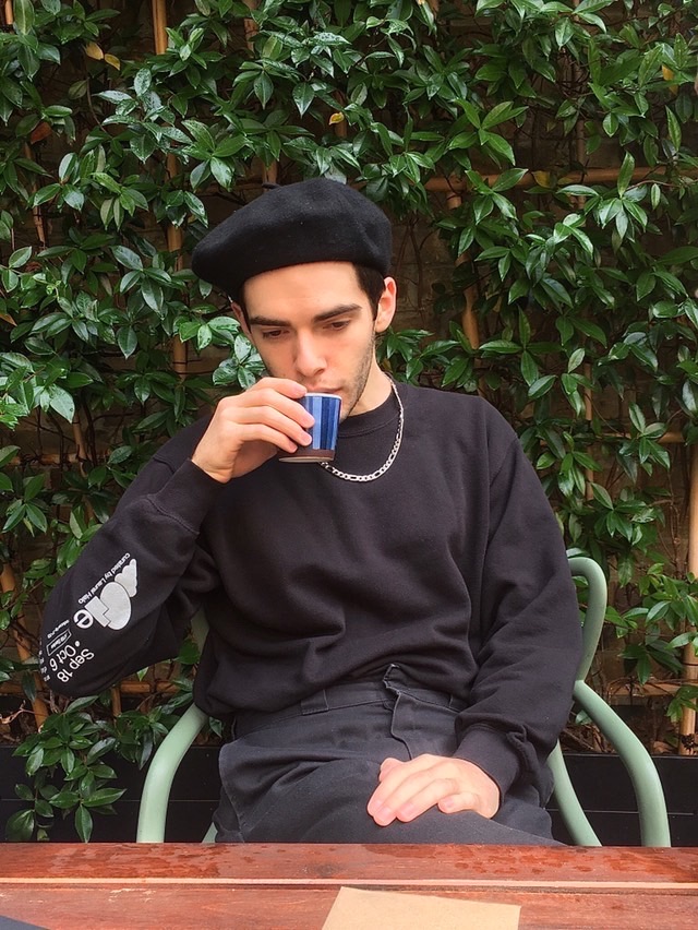 Amos, a young, white man, is wearing an all-black outfit (including beret) and drinking tea in front of a trellis of leafy, climbing plants